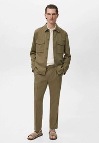 Olijfgroene overhemdjas met twee borstzakken, een witte geribbelde t-shirt eronder, bijpassende broek en bruine sandalen. Gladde textuur, comfortabele pasvorm.