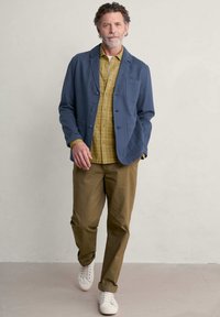 Marineblaue, leichte Jacke mit Reverskragen und Fronttaschen, kombiniert mit einem gelben Karoshemd und khakifarbenen Hosen sowie weißen Sneakers.