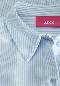 Camicia a righe blu chiaro con colletto classico, chiusura a bottoni sul davanti e etichetta rosa con "JJXX". Tessuto texturizzato con sottili righe.