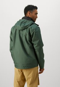 Chaqueta impermeable verde con capucha, con cremallera frontal y puños elásticos, combinada con pantalones en un tono beige claro. Textura suave.