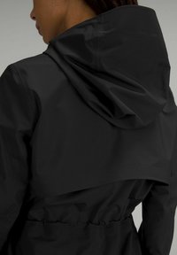 Svart vattentålig jacka med stor huva, åtstramad midja och en slät, glänsande yta. Bakdesignen inkluderar en lagerstruktur med en flikdetalj.