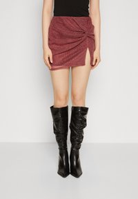 Mujer vestida con una falda corta de textura roja y una abertura frontal, combinada con botas negras holgadas hasta la rodilla, sobre un fondo sencillo.