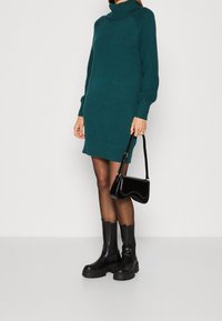 Robe en tricot col roulé bleu canard avec des manches texturées, portée avec des bottines noires et un sac bandoulière noir. Design lisse et ajusté.