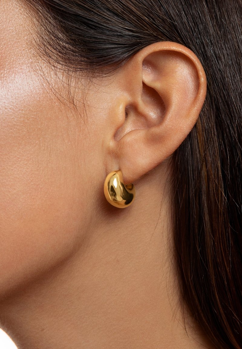 Gros plan sur l’oreille d’une personne portant une petite boucle d’oreille en forme de rein, brillante, de couleur or, avec des cheveux châtain foncé coiffés derrière l’oreille.