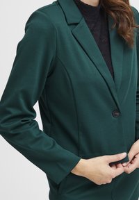 Blazer verde scuro con un design aderente, chiusura con un solo bottone e rever a notched. Realizzato in tessuto liscio, con maniche lunghe e tasche laterali.