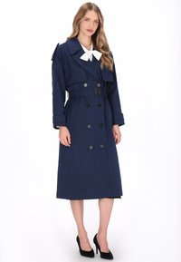 Marineblauwe trenchcoat met dubbele rij knopen, brede kraag, strikdetail en riem; gemaakt van gladde stof met lange mouwen en knielengte ontwerp.