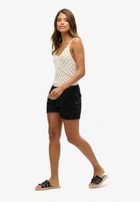 Top de tirantes blanco de ganchillo con escote en V, combinado con pantalones cortos de cargo negros con bolsillos laterales y sandalias de slide negras.