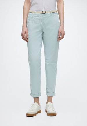 Osoba nosi svetlomodré zrolované chino nohavice s béžovým prepleteným opaskom, biele tenisky so svetlohnedou podrážkou a biely tielkový top.