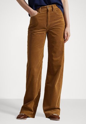 Persona que lleva pantalones de pana marrones de talle alto y pierna ancha con botón y bolsillos delanteros, combinados con zapatos marrones y una camiseta azul marino.