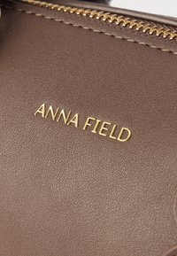 Hnedá kožená kabelka s textúrovaným povrchom, ktorá má na prednej strane zlaté vyrazené logo "ANNA FIELD" a zlatý zips ako doplnok.