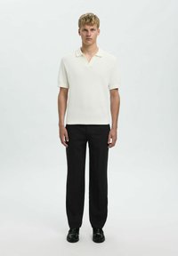 Polo a maniche corte di un leggero colore crema con colletto, abbinato a pantaloni neri e scarpe nere, in piedi su uno sfondo neutro.