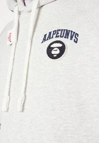 Sweat à capuche gris clair avec cordons blancs et texte bleu marine "AAPEUNVS" au-dessus d'un patch logo visage de singe noir et blanc sur la poitrine.