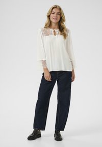 Weißbluse mit Spitze an den Ärmeln und einem Bindedetail. Kombiniert mit lockeren, dunkelblauen Jeans und schwarzen Schuhen. Einfaches, leichtes Material.