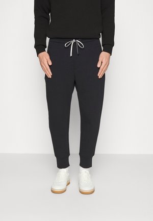 Donkerblauwe joggingbroek met elastische boorden en een trekkoord in de taille; gemaakt van zacht materiaal. Witte sneakers met een beige zool eronder.