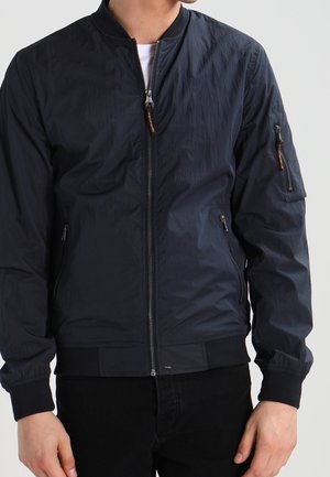 Homme portant une veste bomber bleu marine avec fermeture éclair à l'avant, poches zippées, poignets et ourlet côtelés, associée à un pantalon noir.