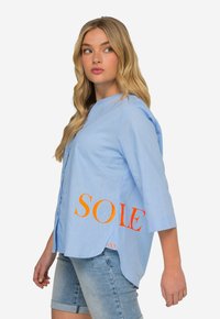 Jasnoniebieska koszula z guzikami i kołnierzykiem mandaryńskim, z rękawami 3/4 oraz pomarańczowym napisem "SOLE" z boku. Miękka, lekka tkanina.