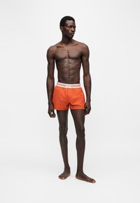 Homme debout pieds nus portant un short de bain orange avec une ceinture blanche à motifs sur un fond blanc uni.