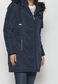 Chaqueta acolchada azul marino con capucha con ribete de piel sintética, cremallera frontal, bolsillos laterales y contrastes negros en las correas.