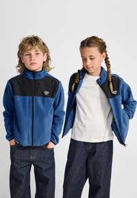 Fleecejas met een blauw lijf, een zwart bovenstuk en een ritssluiting. Heeft zijzakken en een geborduurd logo op de borst.