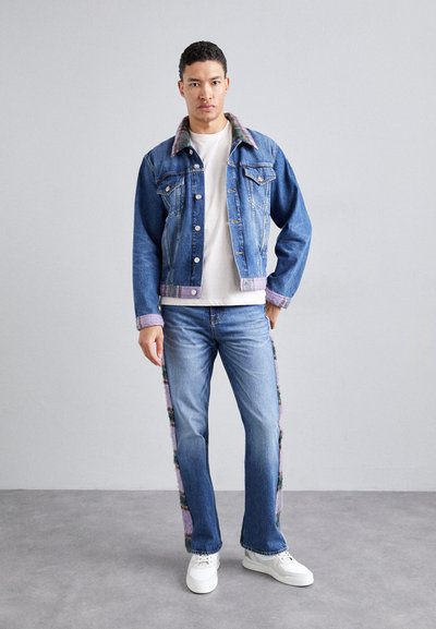 Veste en denim avec un col à motifs violets et verts, t-shirt blanc, jeans bleus avec des panneaux latéraux colorés, et baskets blanches.