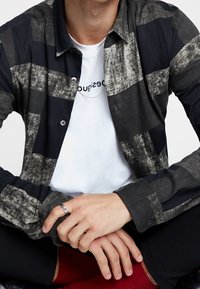 Chemise à manches longues noire et grise avec un motif à rayures, en tissu texturé et poignets boutonnés, portée sur un t-shirt blanc.