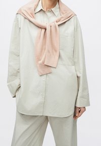 Personne portant une chemise beige ample boutonnée et un pantalon assorti avec un pull rose pâle drapé et noué autour du cou.