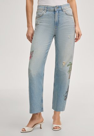 Frau trägt hellblaue High-Waist-Jeans mit Blumen- und Palmenmuster, verwaschenen Details und weißen Sandaletten mit Absatz.