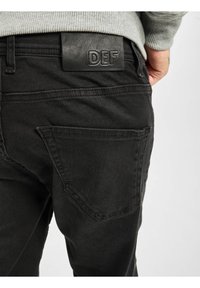 Muž v černých džínách s koženou nášivkou DEF na pasu a šedém žebrovaném svetru, ruka opřená nedaleko boků.