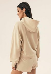 Beżowy, krótki hoodie z kapturem na sznurki, luźny krój, opadające ramiona i ściągacze na nadgarstkach. W zestawie z dopasowanymi szortami w podobnym kolorze.