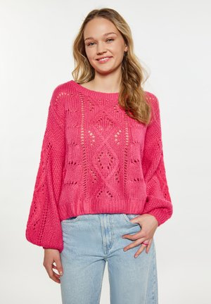 Rosa gestrickter Pullover mit lockerer Passform, der komplizierte Muster, überschnittene Schultern und gerippte Bündchen aufweist. Kombiniert mit hellblauen Jeans.