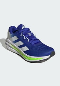Blaues Sport Schuhwerk mit Mesh-Obermaterial, weißen und grünen Akzenten, strukturiertem Sohlen und Schnürsenkeln. Branding auf der Seite und der Zunge sichtbar.