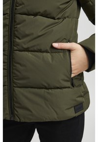 Olivgrön vadderad jacka med quiltad struktur, svart dragkedja och sidoficka. Har en logotyp-tag på den nedre fållen.