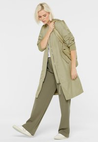 Lange, hellolivgrüne Jacke mit einem Reißverschluss und gerippten Bündchen. Kombiniert mit passenden, weit geschnittenen Hosen und weißen Sneakers, vor einem schlichten Hintergrund.