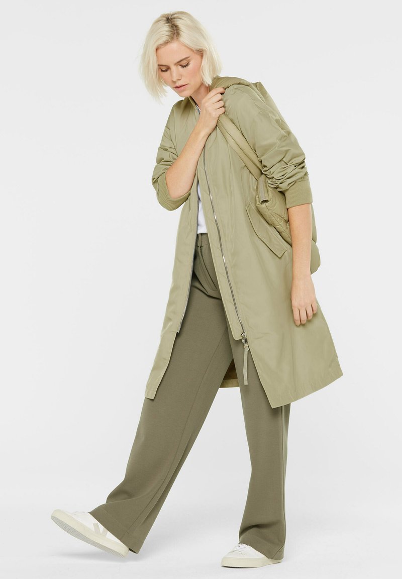 Lange, hellolivgrüne Jacke mit einem Reißverschluss und gerippten Bündchen. Kombiniert mit passenden, weit geschnittenen Hosen und weißen Sneakers, vor einem schlichten Hintergrund.