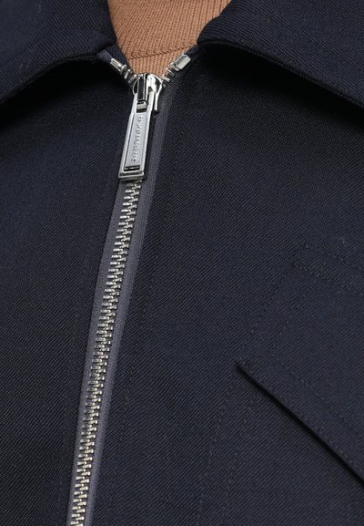 Chaqueta de color azul marino con un brillante zipper plateado, que incluye un cuello y un bolsillo en el pecho. Tejido texturizado con costuras visibles.