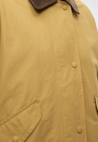 Manteau kaki avec un col en velours côtelé marron, doté de boutons-pression, de double surpiqûre et de poches avant à bords arrondis.