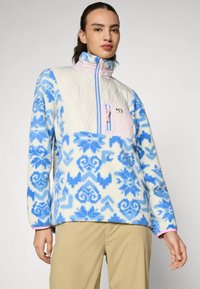Sudadera de forro polar con la parte superior acolchada en crema y la sección inferior azul con patrón. Cuenta con un bolsillo con cremallera rosa y un cuello alto. Textura suave.