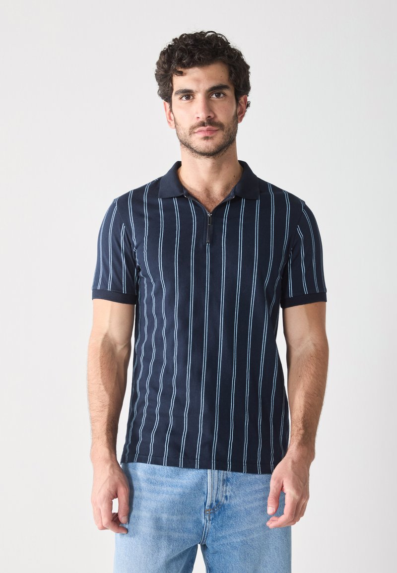 Polo navy avec des rayures verticales bleu clair, manches courtes et col zippé. Tissu lisse avec une coupe classique. Porté avec un jean bleu clair.