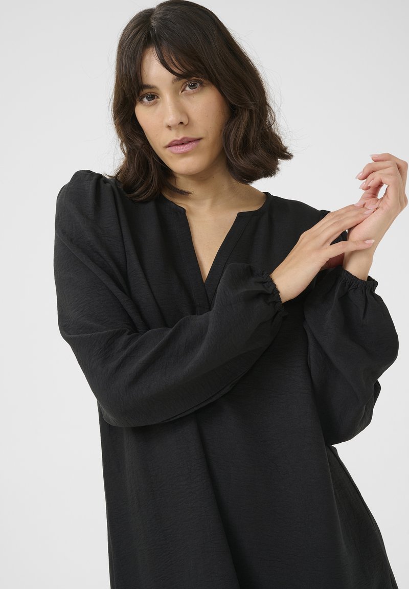Femme aux cheveux foncés mi-longs portant un chemisier noir à manches longues, touchant son poignet sur un fond clair uni.