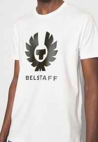 Camiseta de algodón blanca con un gran logo de águila negra y el texto "BELSTAFF" en la parte inferior. Mangas cortas y cuello redondo.