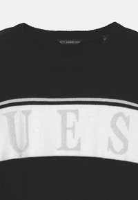 Mustapusero, jossa on pyöreä kaula-aukko, valko-harmaa raitakuosi ja "GUESS" -logo, joka on selvästi näkyvissä harmailla kirjaimilla.