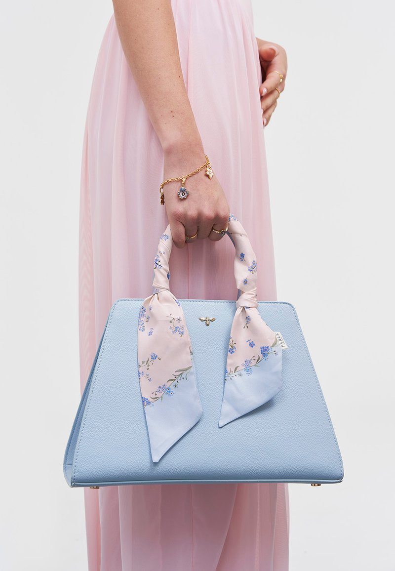 Vrouw in roze jurk die een lichtblauwe handtas vasthoudt met handvatten omwikkeld met een bloemen sjaal en een gouden bijenembleem, draagt gouden ringen en een bedelarmband.