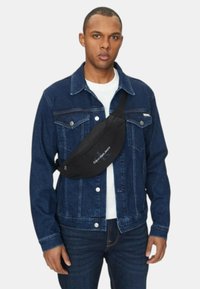 Zwarte stoffen heuptas gedragen over een blauwe spijkerjack, met een subtiele logo, gebogen vorm en verstelbare band voor een veilige pasvorm.