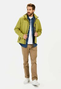 Grøn vandtæt jakke med aftagelig hætte, frontlynlås og to sidelommer, lagdelt over en denimskjorte og beige bukser. Hvide sko.