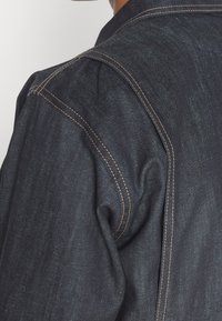 Primer plano de una chaqueta de mezclilla azul oscura que muestra un detallado pespunte beige a lo largo del hombro y la costura trasera.