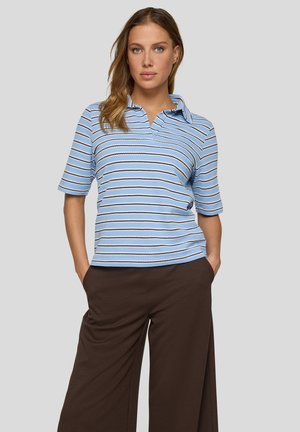 Frau trägt ein hellblau gestreiftes kurzärmliges Poloshirt und lockere braune Hose, steht mit den Händen in den Taschen vor einem schlichten Hintergrund.