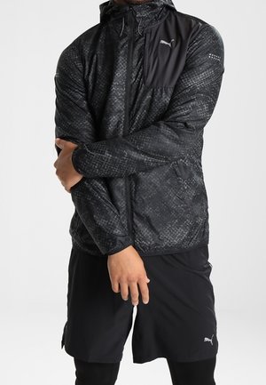 Uomo con giacca a vento Puma nera con motivo a fantasia e cappuccio e pantaloncini Puma neri, con le braccia incrociate davanti a uno sfondo bianco.