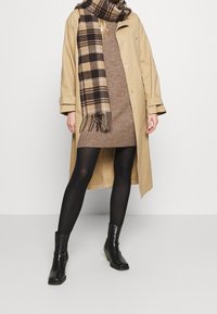 Trench-coat beige avec boutons argentés, robe en maille marron, collants noirs et bottines. Accessoirisé d'une écharpe à carreaux marron avec franges.