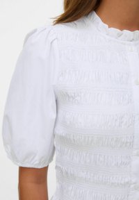 Blouse blanche avec un design smocké texturé, des manches bouffantes et un col froncé, complétée par des boutons sur le devant.