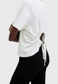 Bílý tričko s krátkým rukávem a staženým designem na zádech, který obsahuje kruhový otvor a nastavitelné tkaničky. Hladká bavlněná tkanina, volný střih.